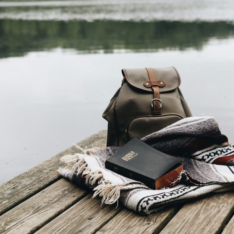 A backpack next to a blanket and a book on a wooden dock by a calm lake.