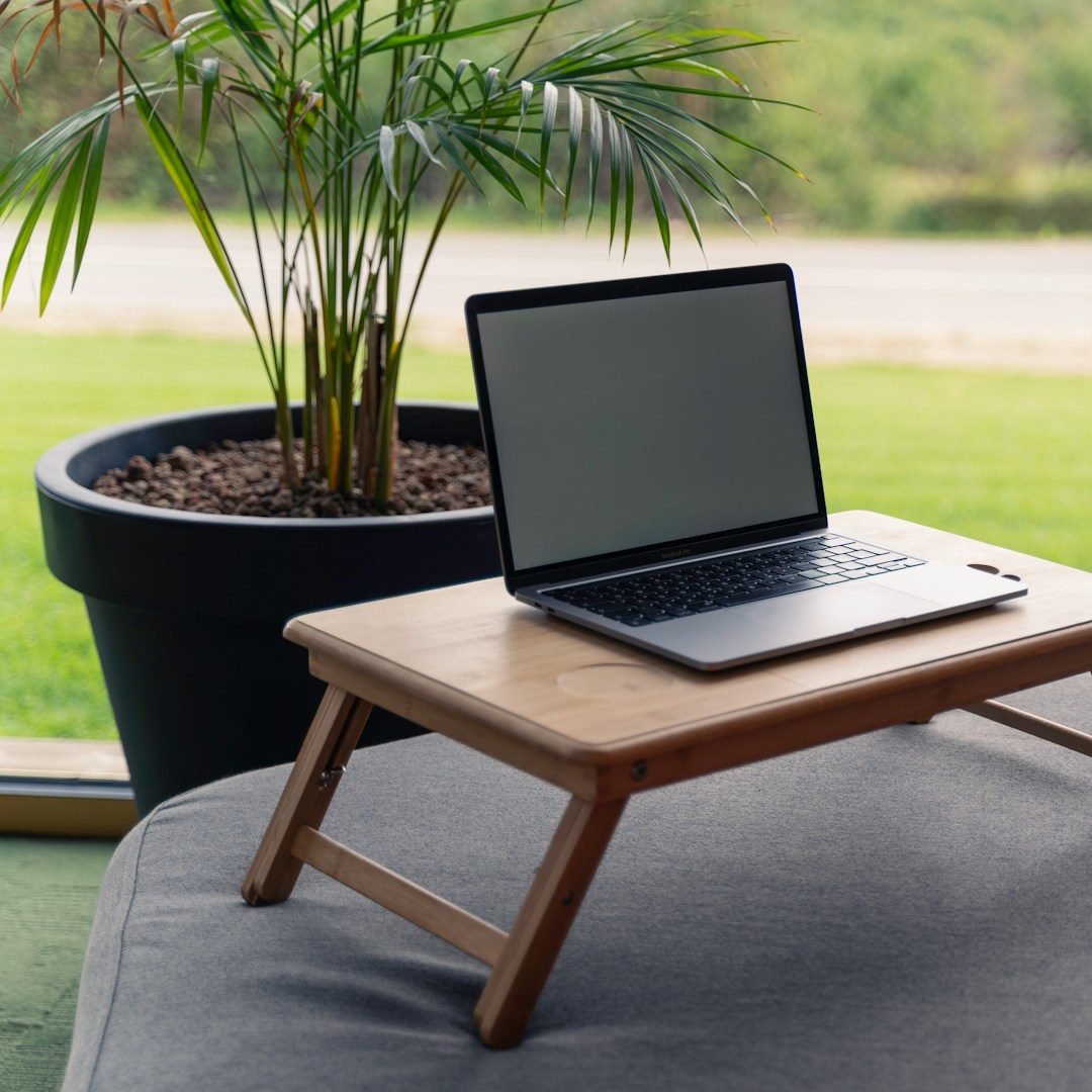 A laptop on a wooden table next to a large potted plant by a window.