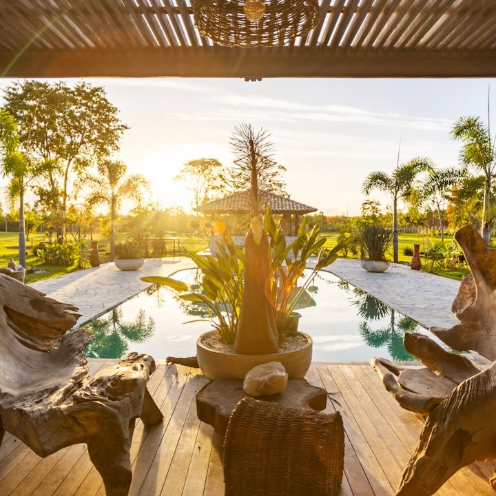 Sunlit patio with pool, tropical plants, and wooden furniture.