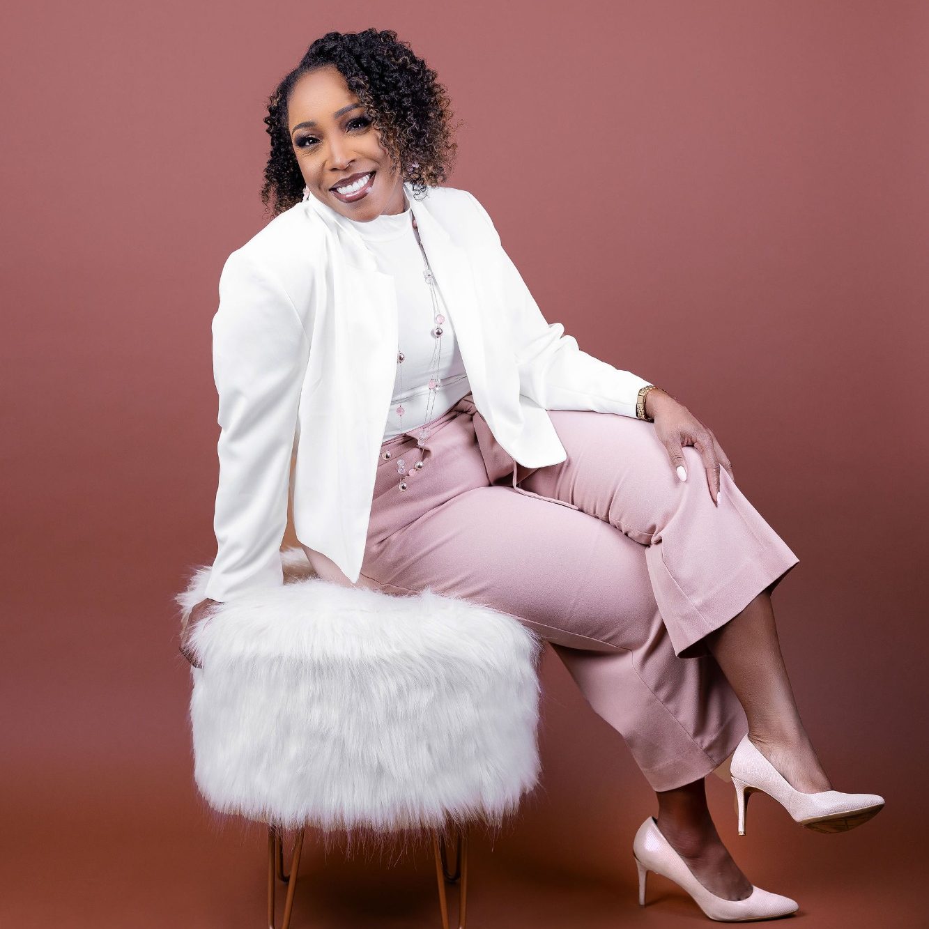 A woman in a white jacket and pink pants sits confidently on a fluffy stool.